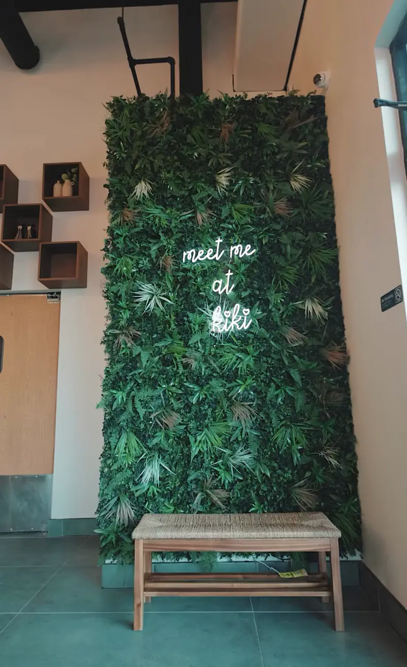 Green living wall with the neon sign meet me at kiki and a wood bench inside Kiki Kafé coffee shop in Clinton Township
