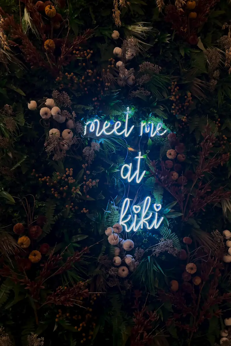 Neon sign that reads meet me at kiki on a dark floral wall